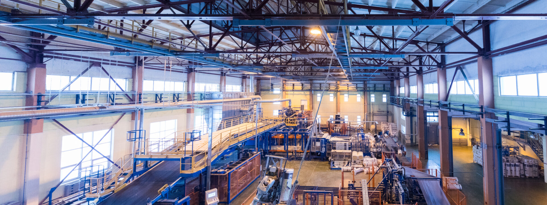 Interior of a large industrial factory with metal beams, machinery, conveyor belts, and stacked materials under bright lighting. The space appears spacious and well-organized, with large windows letting in natural light.