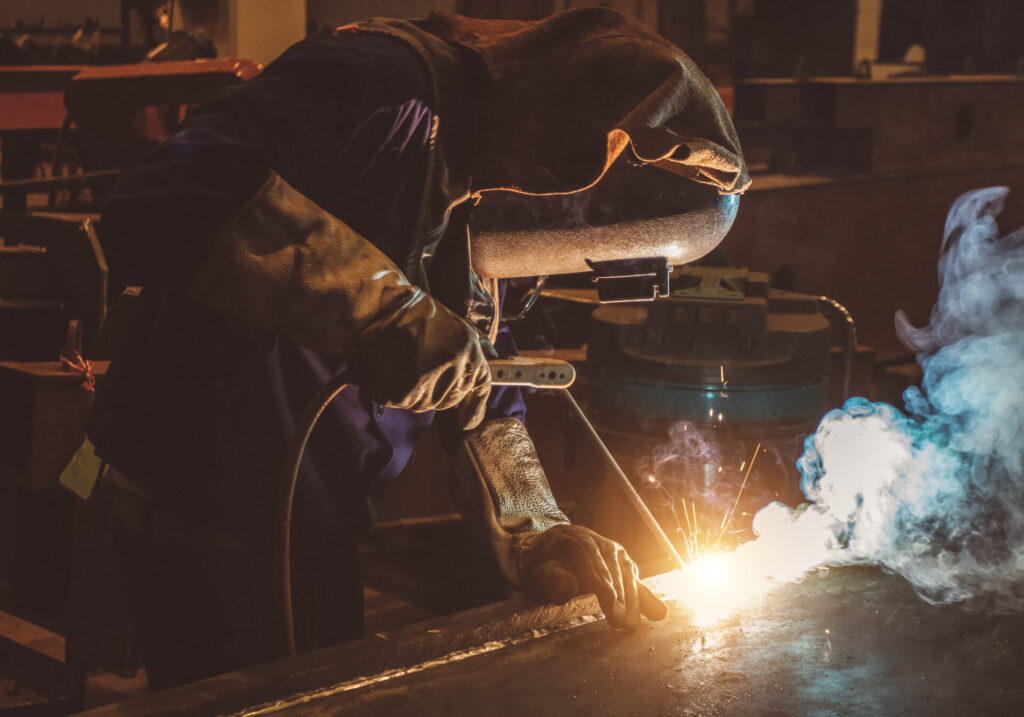 A person wearing protective gloves and a welding helmet uses a welding tool, creating bright sparks and smoke while working on a metal surface in an industrial workshop.