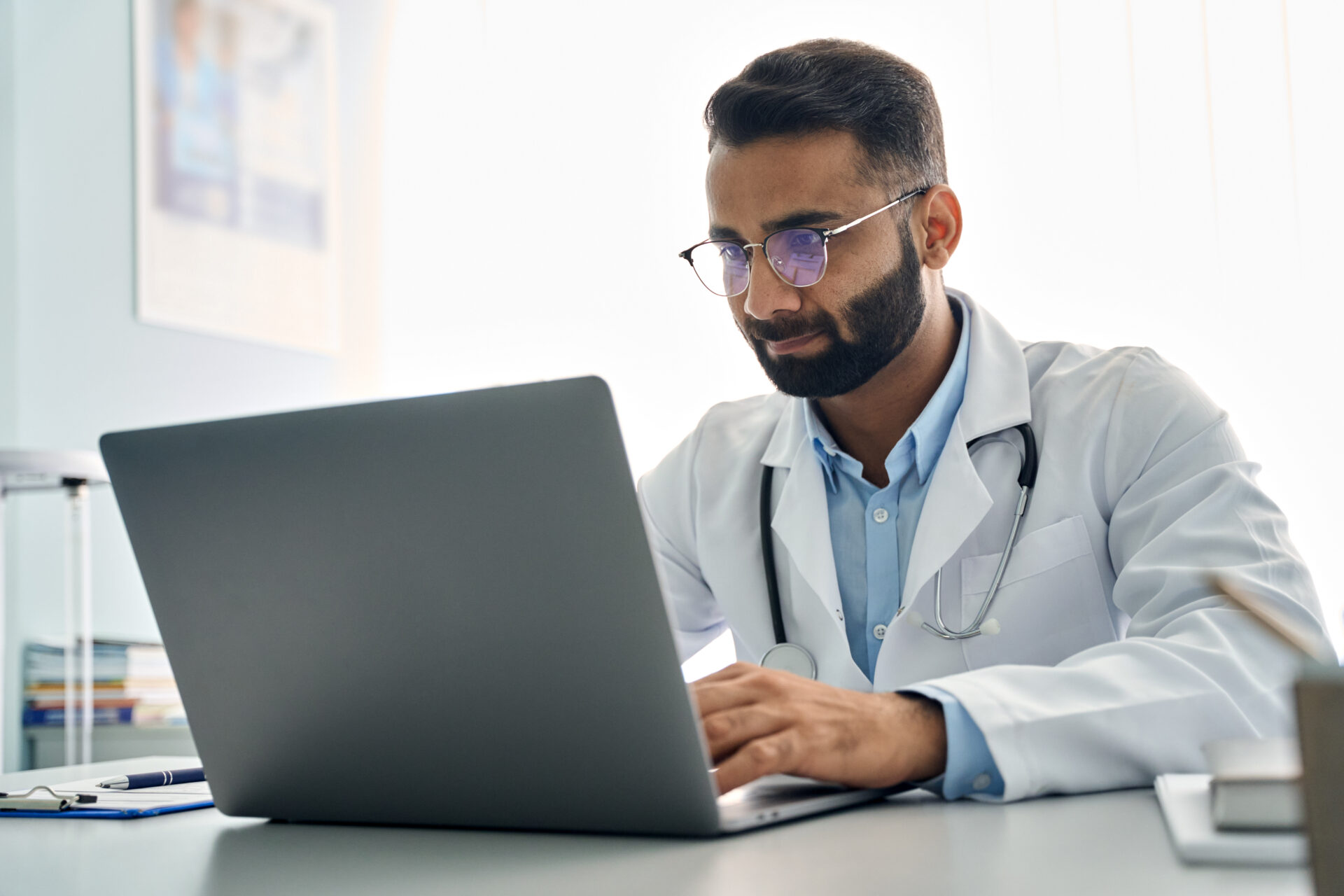 A bearded male doctor with glasses in a white coat and stethoscope works on a laptop at his desk in a bright office.