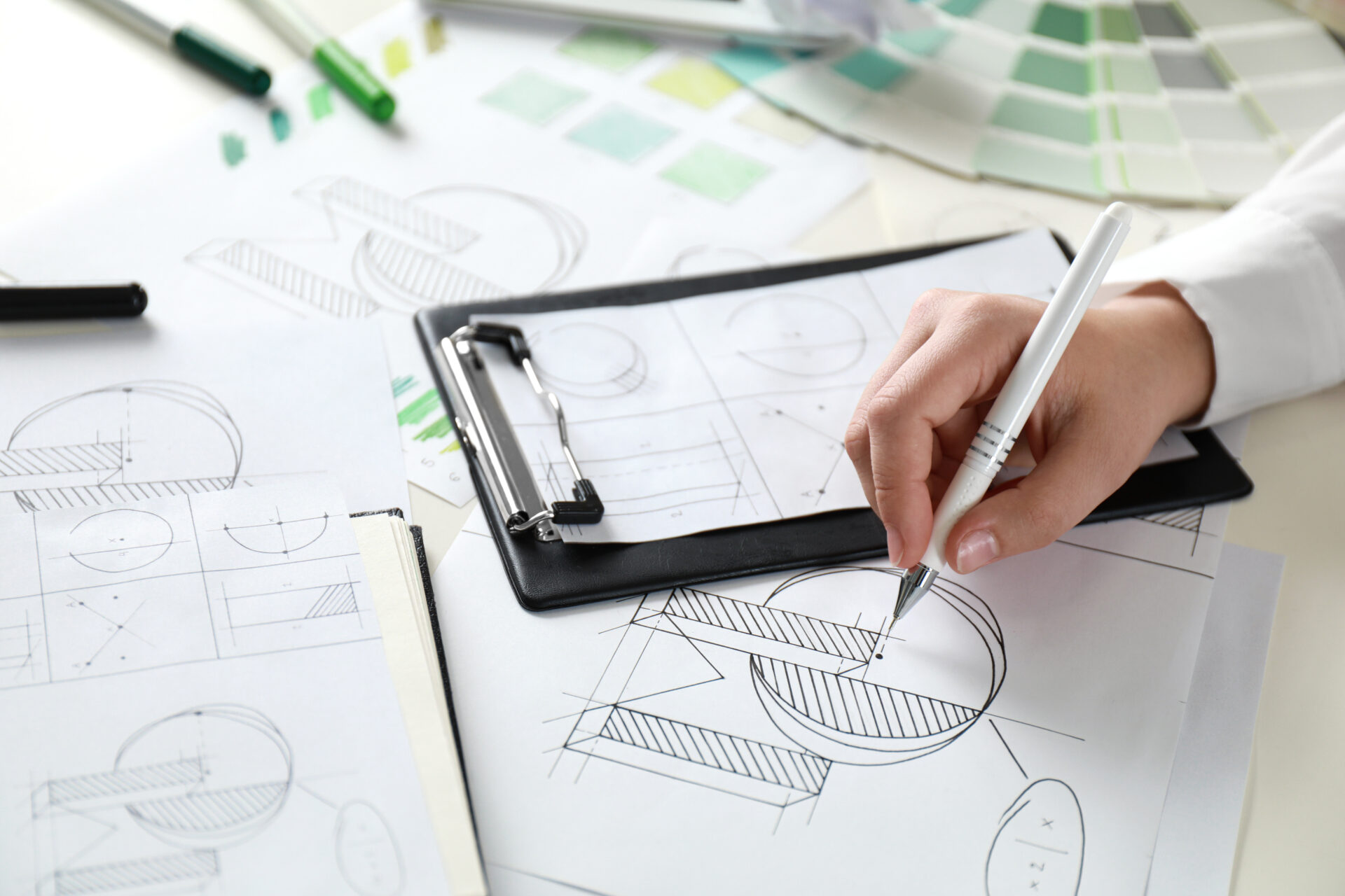 A person sketches geometric logo designs on paper with a pen. Various design drafts, color swatches, and drawing tools are scattered on the desk, indicating a creative design process.