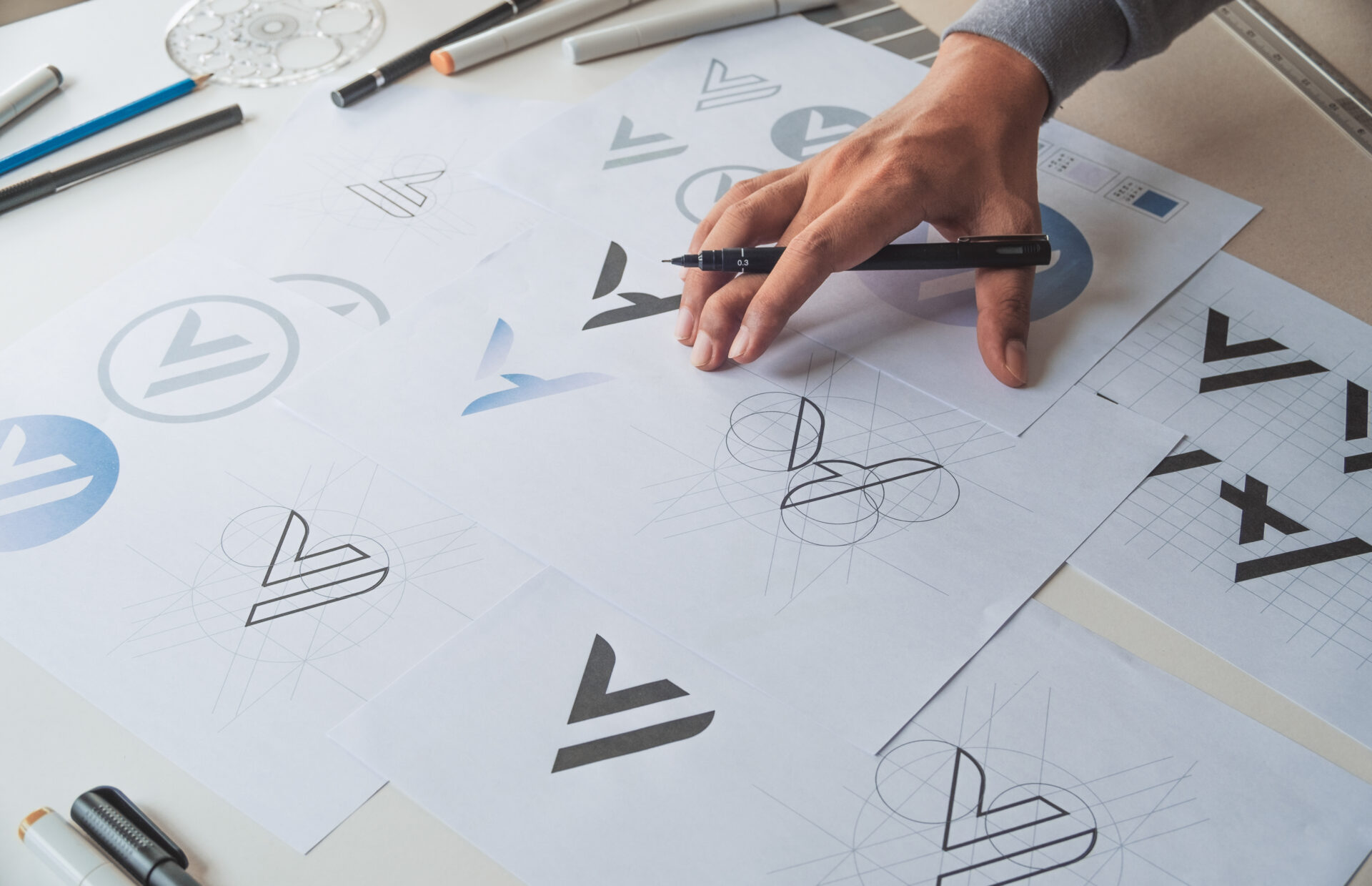 A person’s hand draws logo designs on white paper, surrounded by sheets featuring geometric sketches and variations of a stylized V symbol, along with pens, pencils, and design tools scattered on the table.