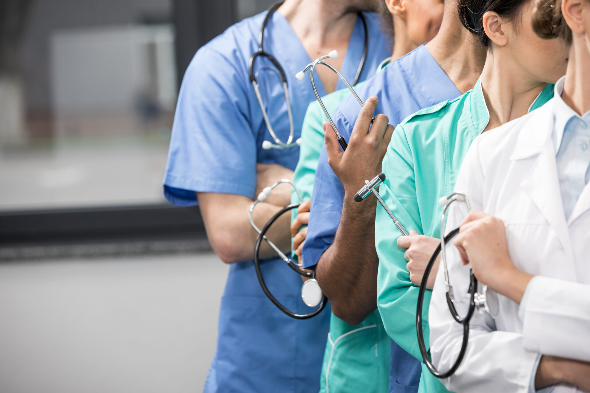 A diverse team of healthcare professionals in scrubs and lab coats stand side by side with stethoscopes, faces partly out of view.