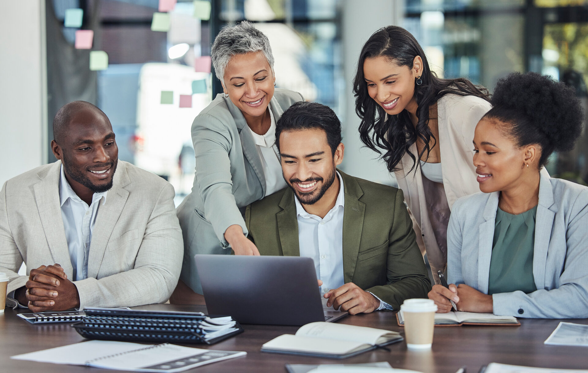 Five diverse banking professionals smile and gather around a laptop in a modern office, collaborating on a new logo design with notebooks, documents, and coffee nearby.