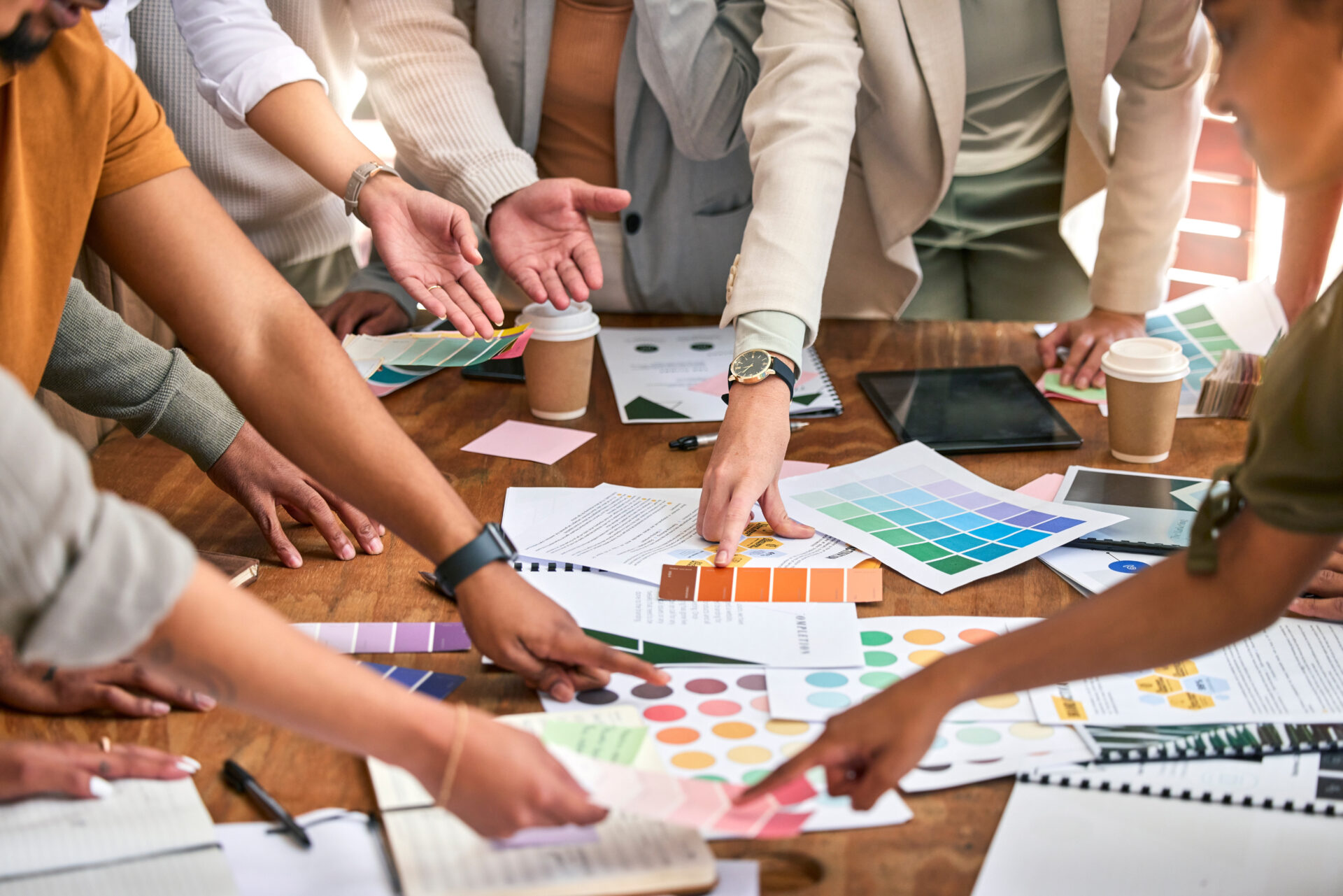 A group of people gather around a table covered with color swatches, design samples, papers, and coffee cups, collaborating and pointing at different materials during a logo design update meeting.