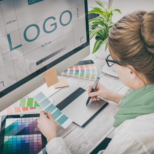 A woman designs a logo on her computer at a desk with plants, color swatches, and design tools, using a graphics tablet.