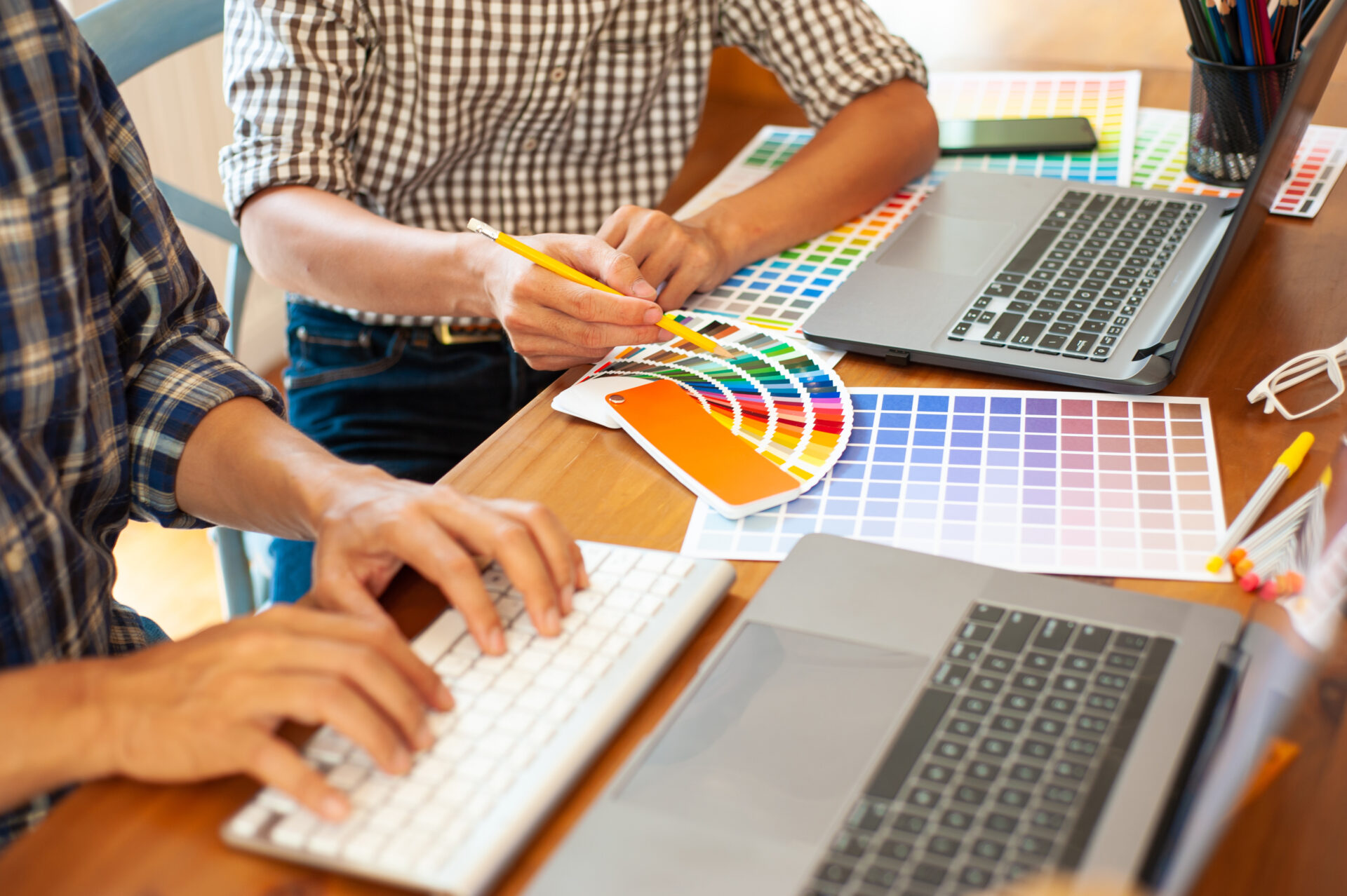Two people working at a desk with laptops, color swatches, and a keyboard, discussing and selecting colors from a palette for a logo design update.