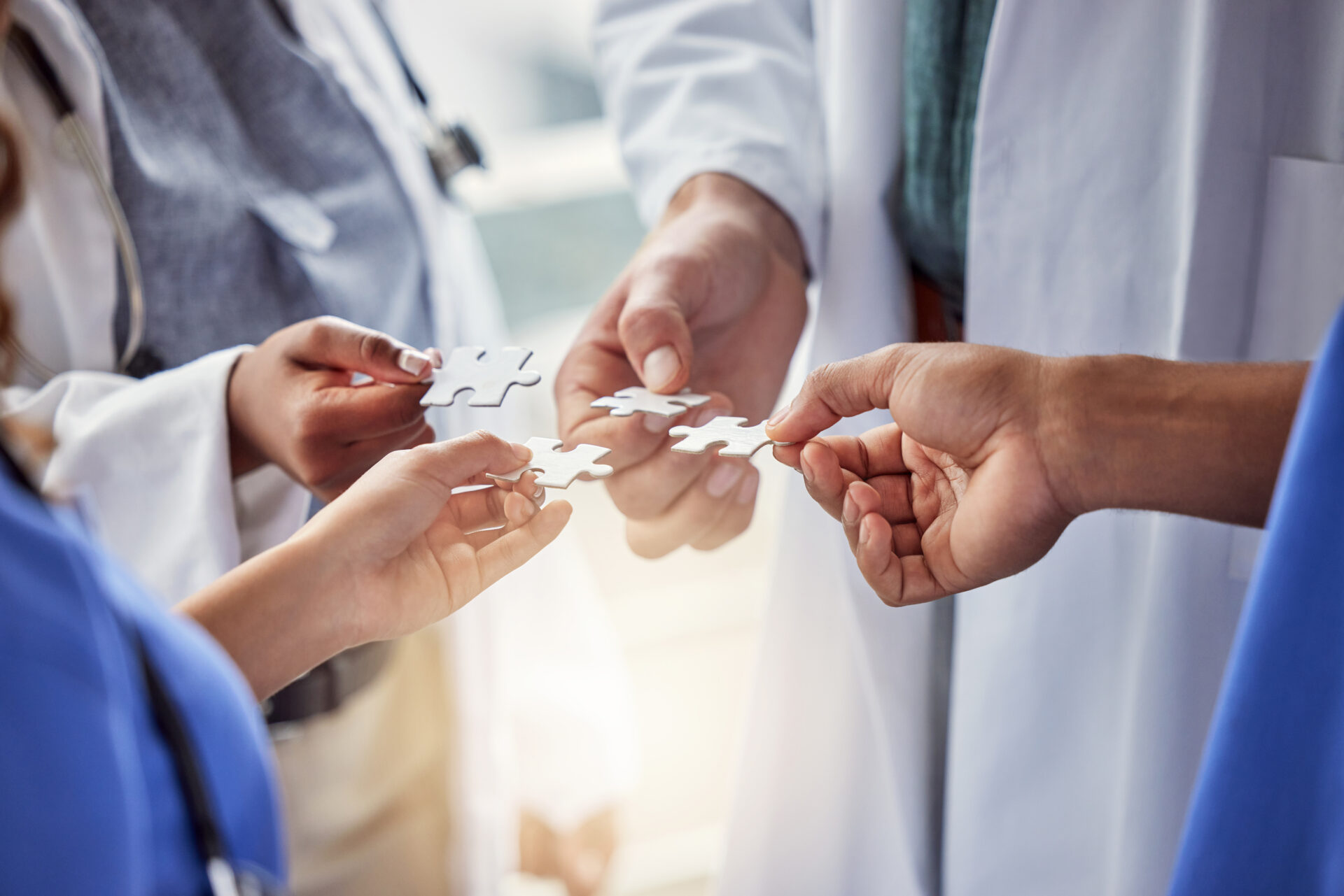 Four people in lab coats and scrubs connect white puzzle pieces, representing teamwork and collaboration in a healthcare setting.