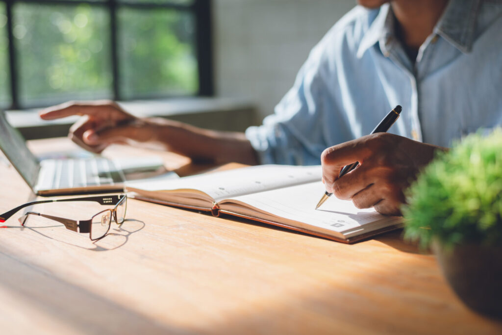 A business person at a wooden desk writes in a notebook and points at a laptop. Eyeglasses, a small plant, and sunlight through a window nearby.