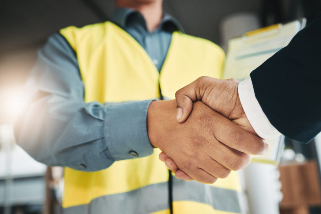 A person in a yellow safety vest shakes hands with someone in a suit jacket, indicating partnership, with a clipboard in the background.