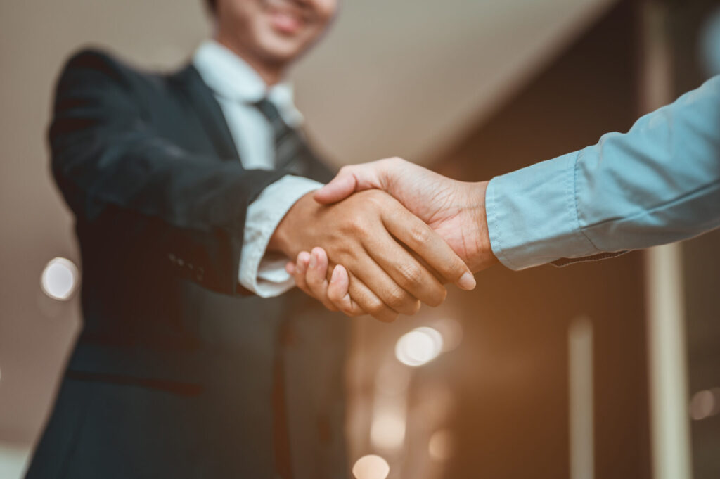 Close-up of two people shaking hands, one in a dark suit and one in a light shirt, indicating business partnership, blurred background.