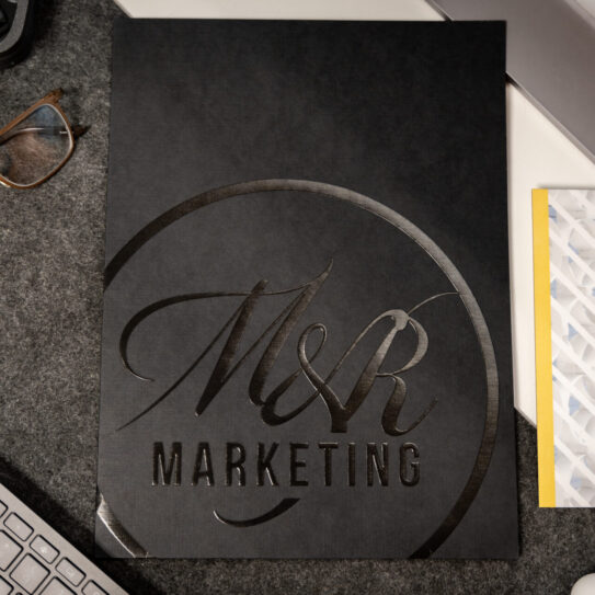A black folder embossed with M&R Marketing lies on a grey desk mat next to a computer keyboard, glasses, and a patterned envelope.