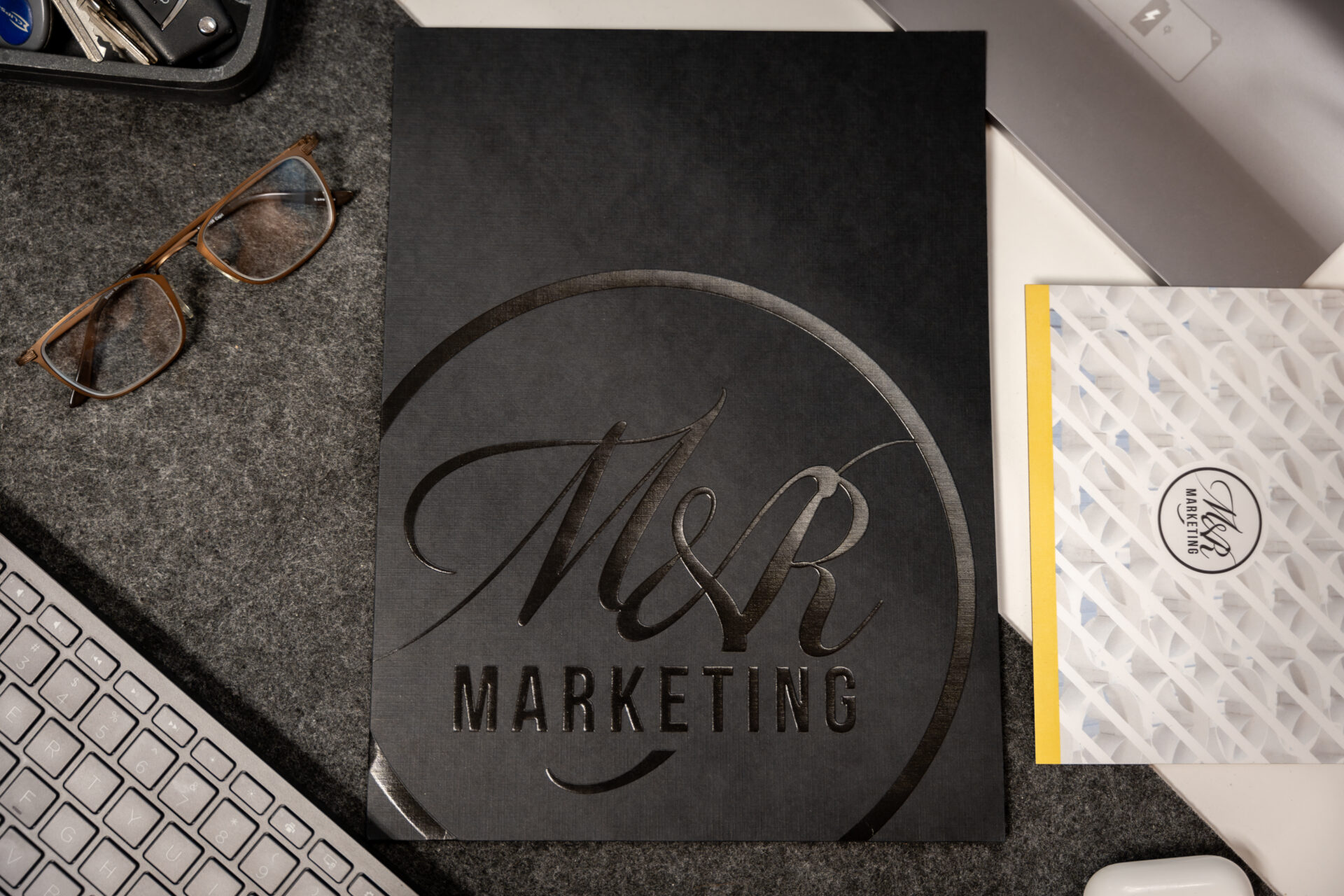 A black folder embossed with M&R Marketing lies on a grey desk mat next to a computer keyboard, glasses, and a patterned envelope.