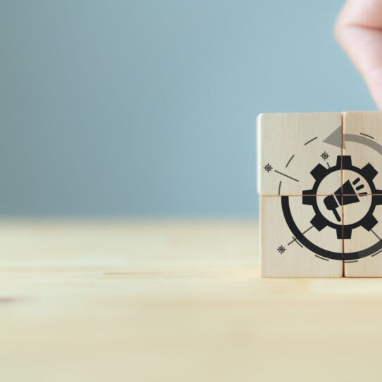 A hand arranges four wooden blocks on a light surface, forming a gear with a megaphone symbol, suggesting marketing automation and tracking