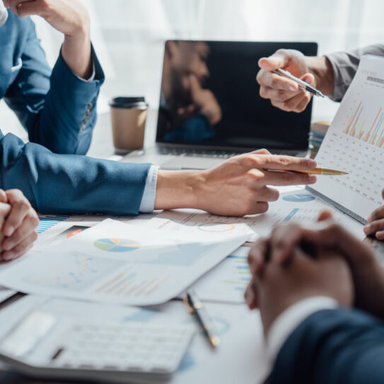 Close-up of business people in suits around a table reviewing charts, one pointing to data on a clipboard with a laptop in the background.