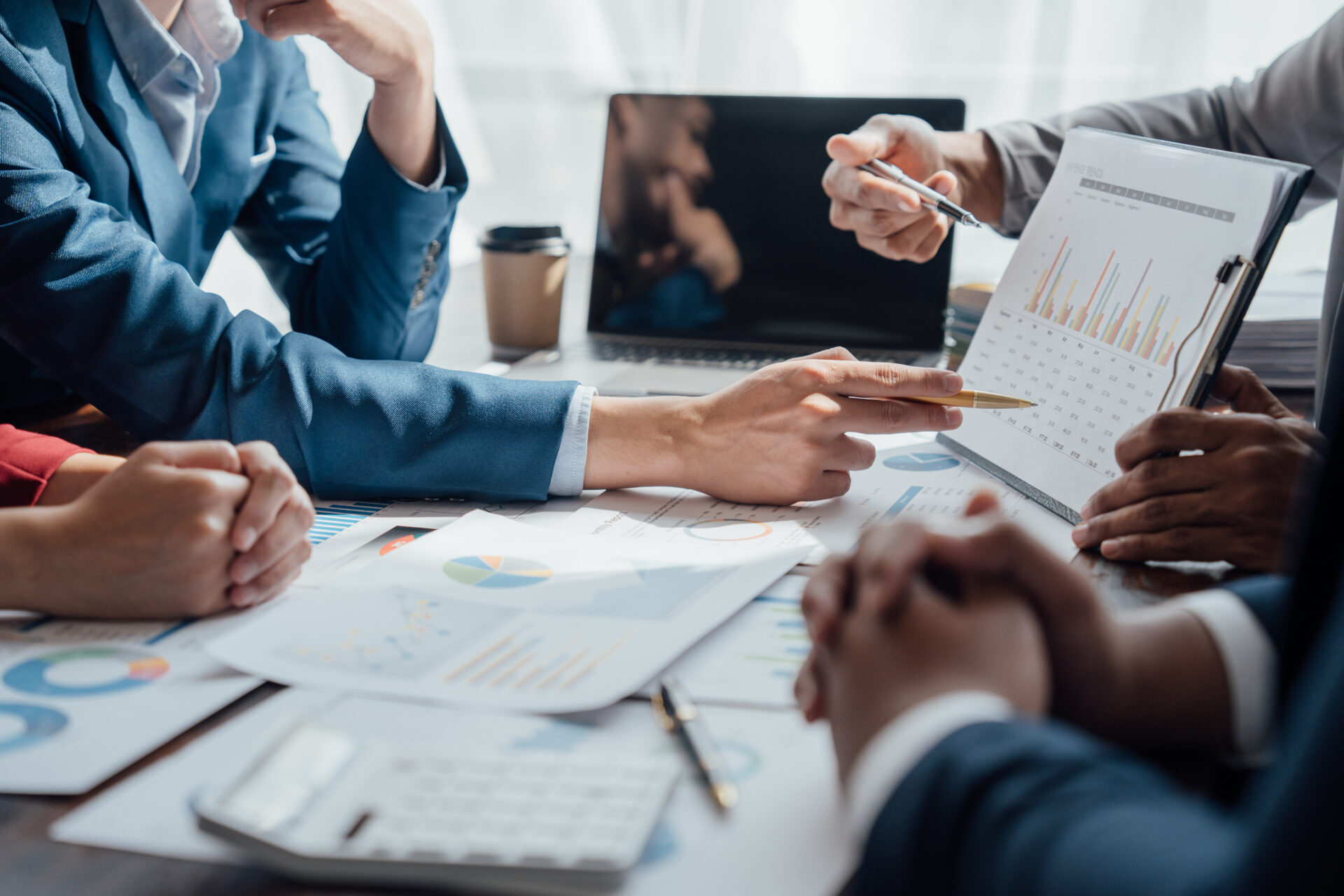 Close-up of business people in suits around a table reviewing charts, one pointing to data on a clipboard with a laptop in the background.