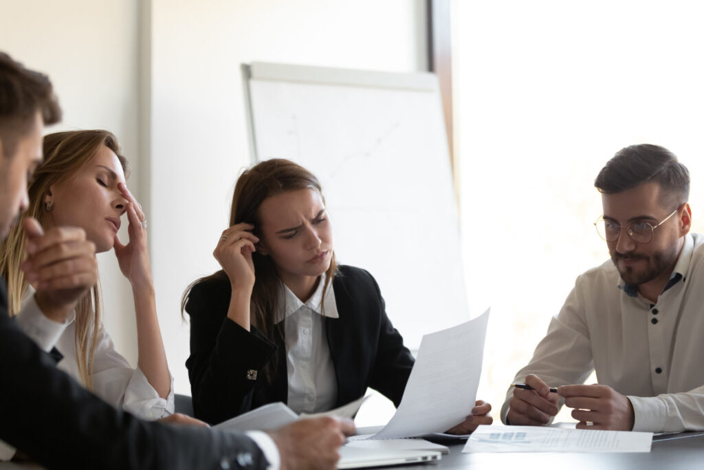 Four business professionals sit around a table with documents, looking stressed as they review paperwork in a bright office.