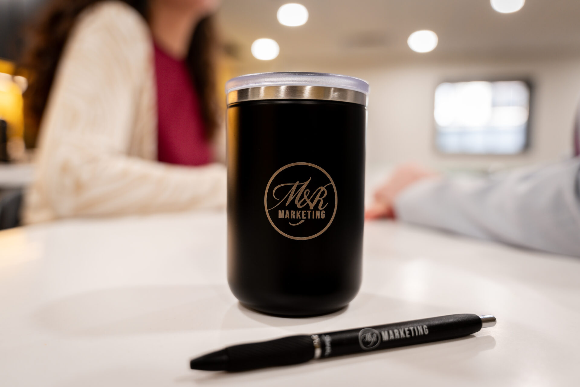A black tumbler with the M&R Marketing logo sits on a white table by a branded pen, with two people talking in a bright room behind.