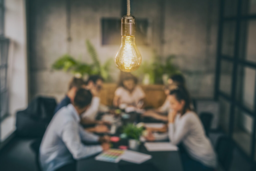 A glowing light bulb hangs above a blurred group meeting around an office table, surrounded by plants and modern decor.