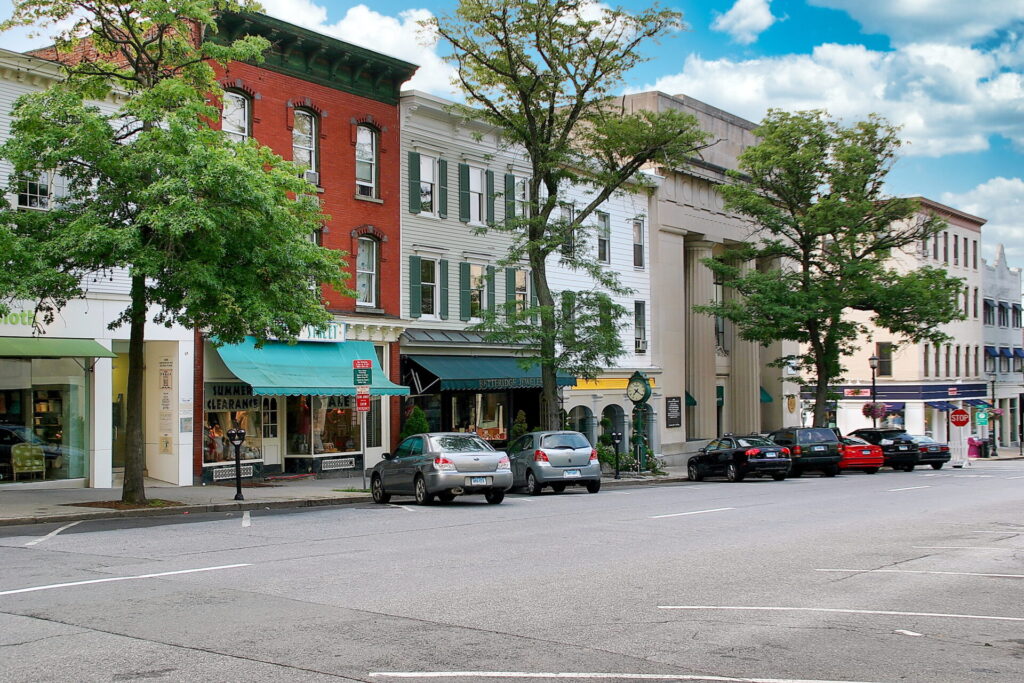 Historic storefronts with awnings and large windows line a tree-lined street. Parked cars sit by the curb on a sunny day.