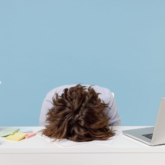 A person sits at a white desk with their head down on papers, looking stressed. The desk has a laptop, lamp, plant, and sticky notes.