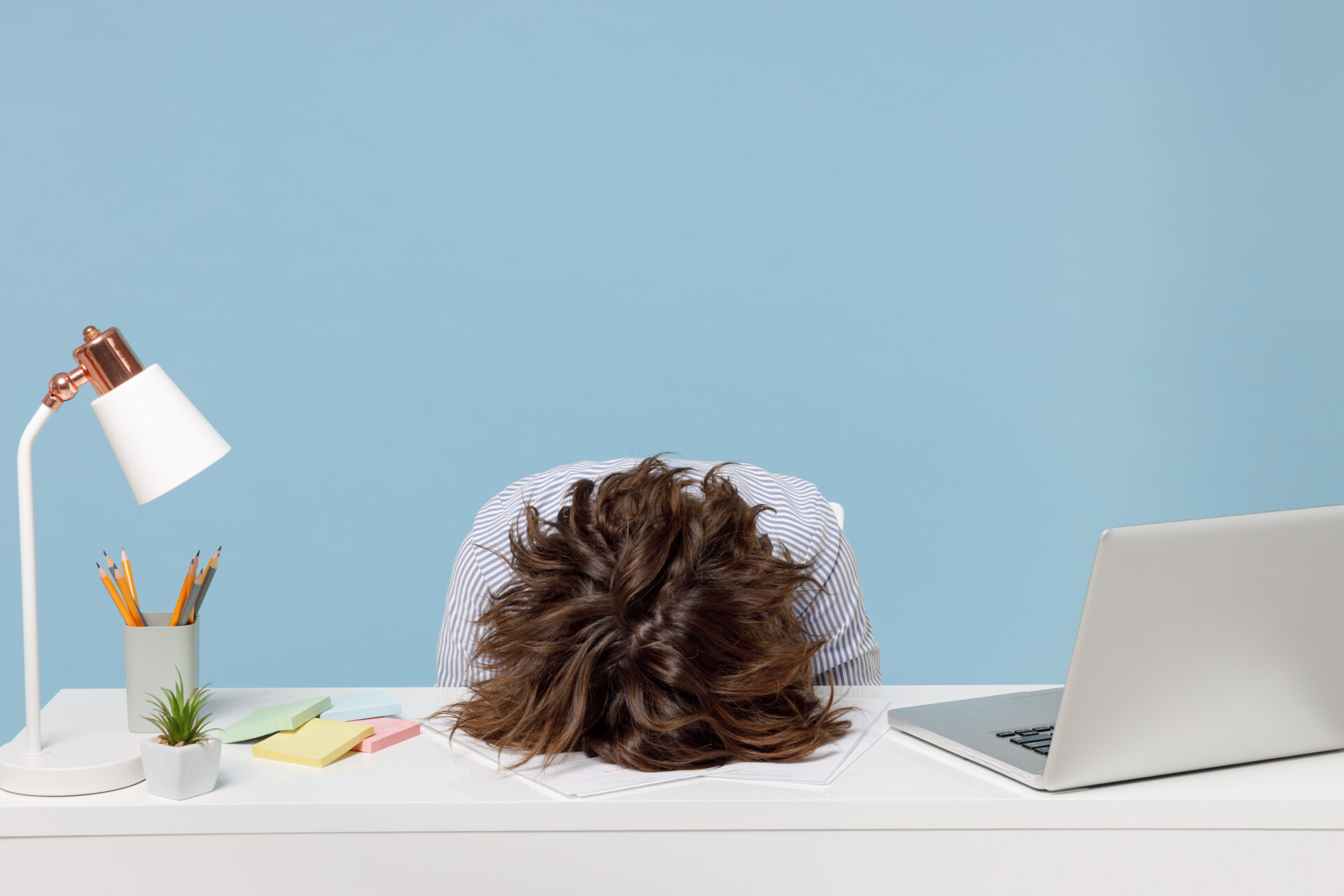 A person sits at a white desk with their head down on papers, looking stressed. The desk has a laptop, lamp, plant, and sticky notes.