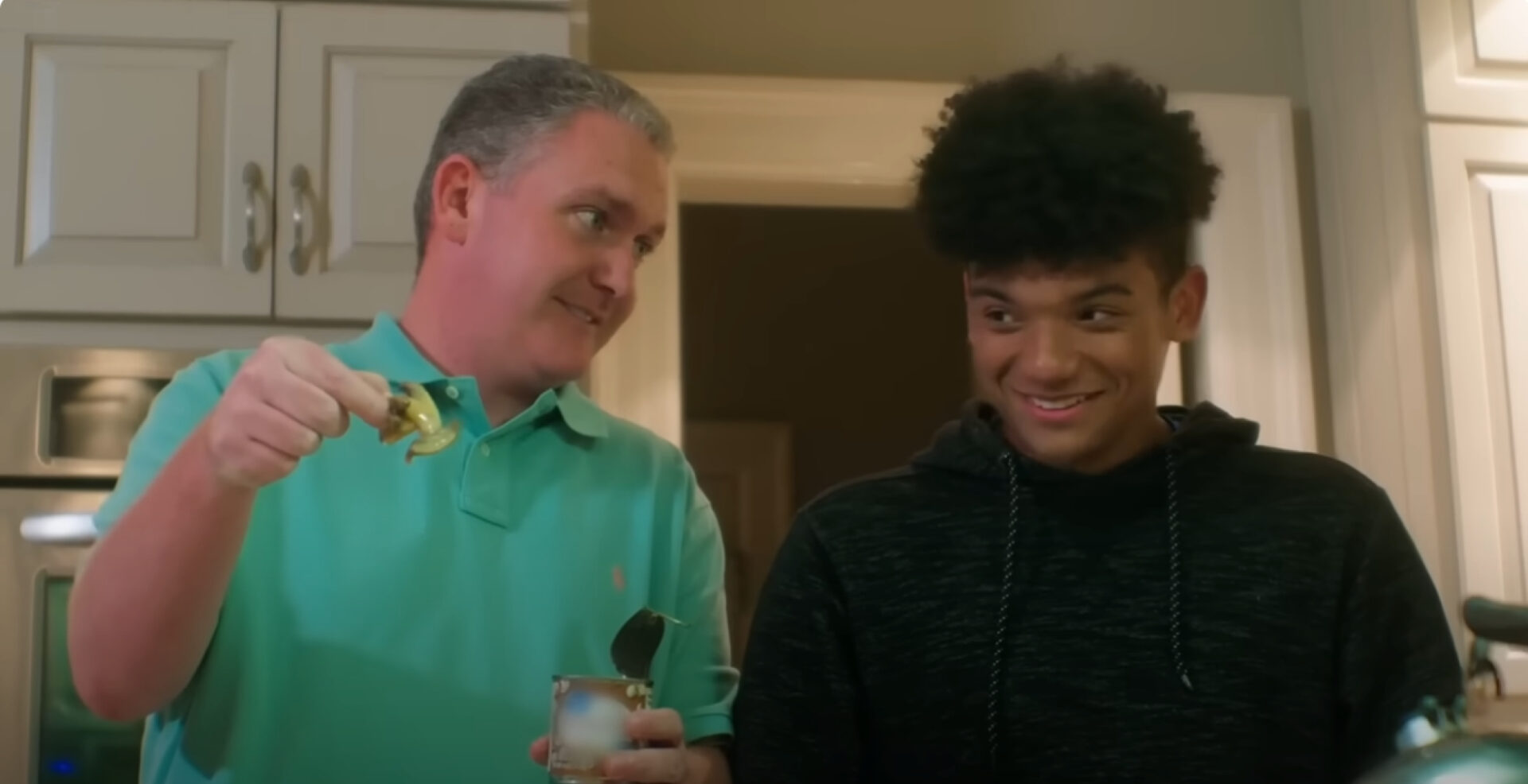 A man in a green polo shirt with a can and can opener smiles at a young man in a black hoodie. They're in a kitchen with white cabinets.
