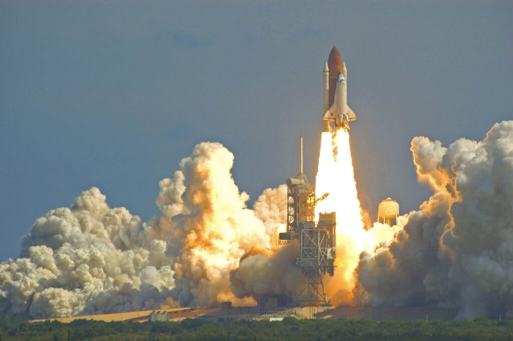 A space shuttle blasts off vertically, leaving fiery exhaust and heavy smoke behind, with a launch pad and green plants in front.