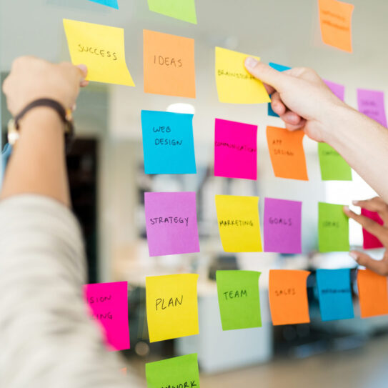 Two people arrange colorful sticky notes with words like SUCCESS, IDEAS, WEB DESIGN, and STRATEGY on a glass wall in an office.