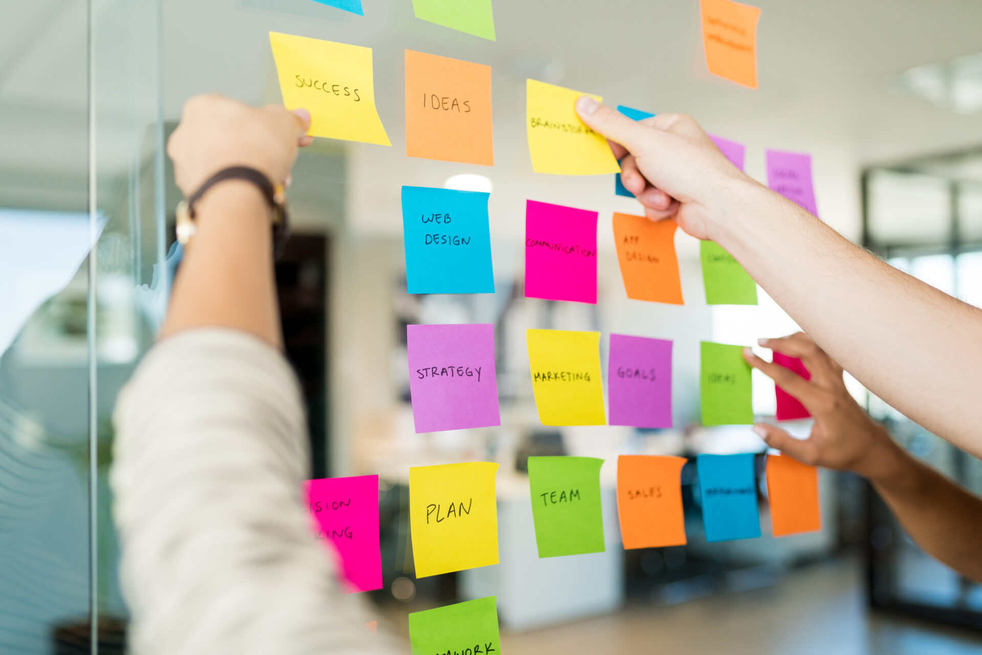 Two people arrange colorful sticky notes with words like SUCCESS, IDEAS, WEB DESIGN, and STRATEGY on a glass wall in an office.