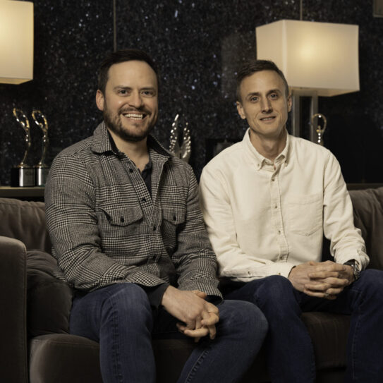 Two men smile while sitting on a brown sofa in a modern room with dark walls, two large lamps, and several awards in the background.