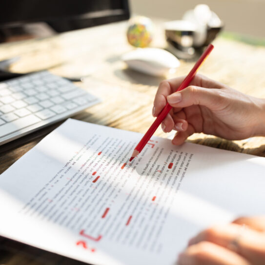 A person marks corrections with a red pencil on a printed document at a sunlit desk with a keyboard and monitor.