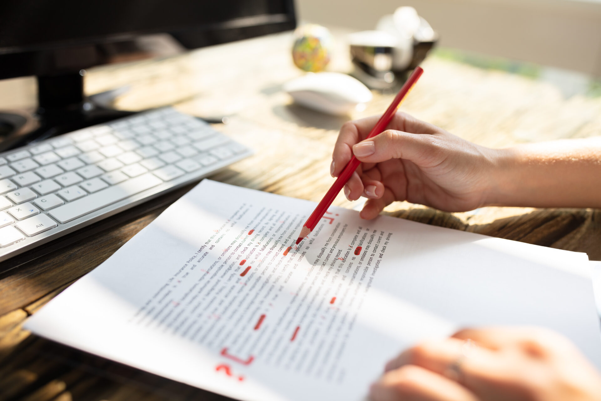 A person marks corrections with a red pencil on a printed document at a sunlit desk with a keyboard and monitor.