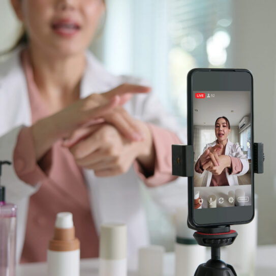 A woman in a white coat applies skincare to her hand during a livestream, filmed by a smartphone. Skincare bottles are on the table.