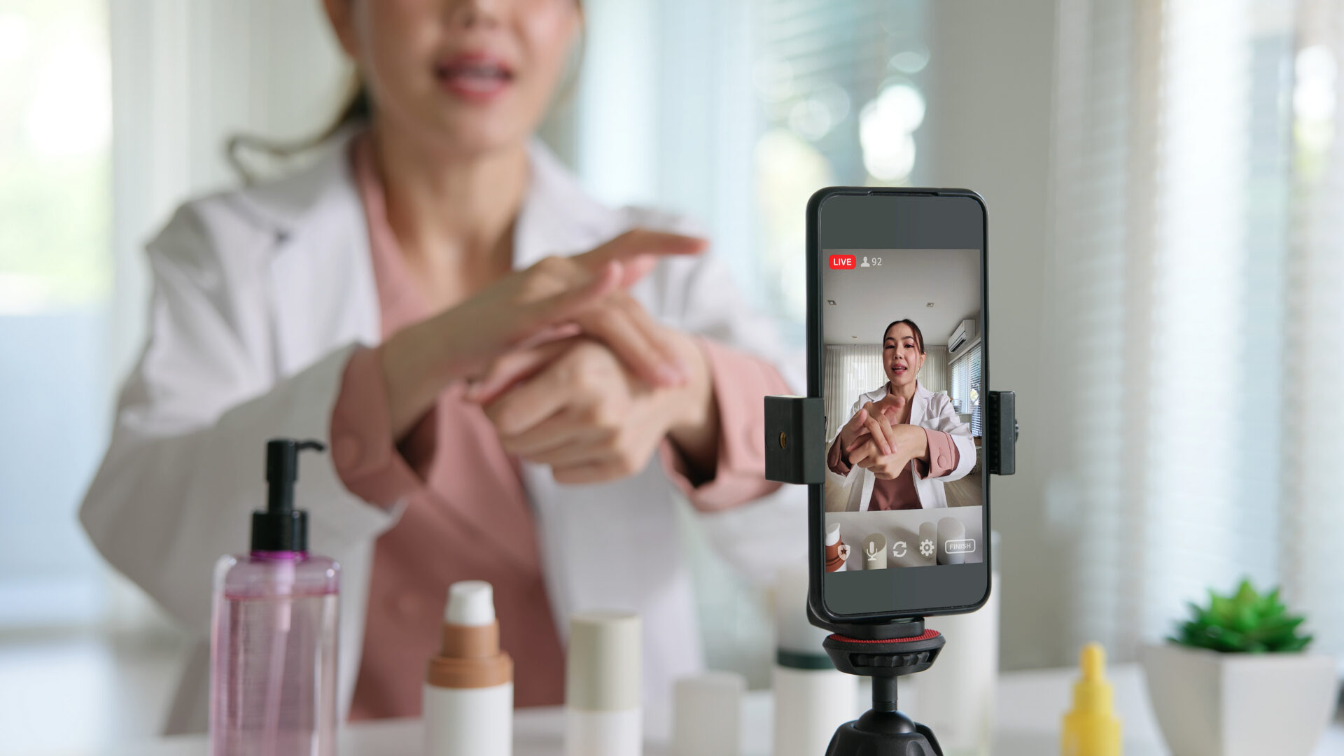 A woman in a white coat applies skincare to her hand during a livestream, filmed by a smartphone. Skincare bottles are on the table.