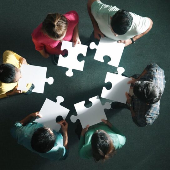 Seven people stand in a circle on a green carpet, viewed from above, each holding a blank puzzle piece, symbolizing teamwork.