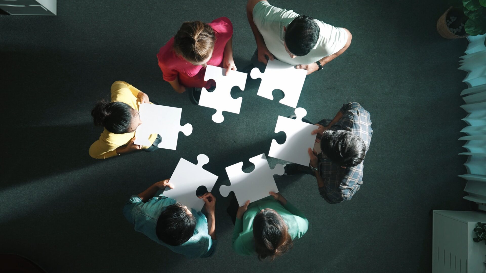 Seven people stand in a circle on a green carpet, viewed from above, each holding a blank puzzle piece, symbolizing teamwork.