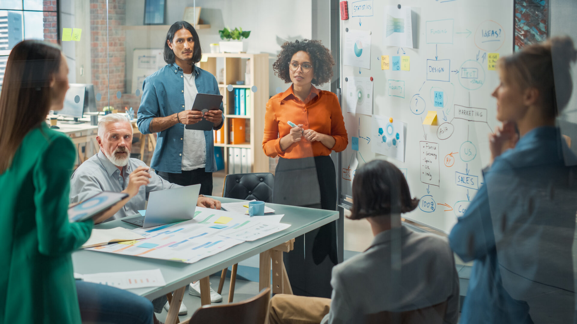 Coworkers in a modern office meet; some sit at a table with papers and a laptop, others stand by a whiteboard with notes and diagrams.