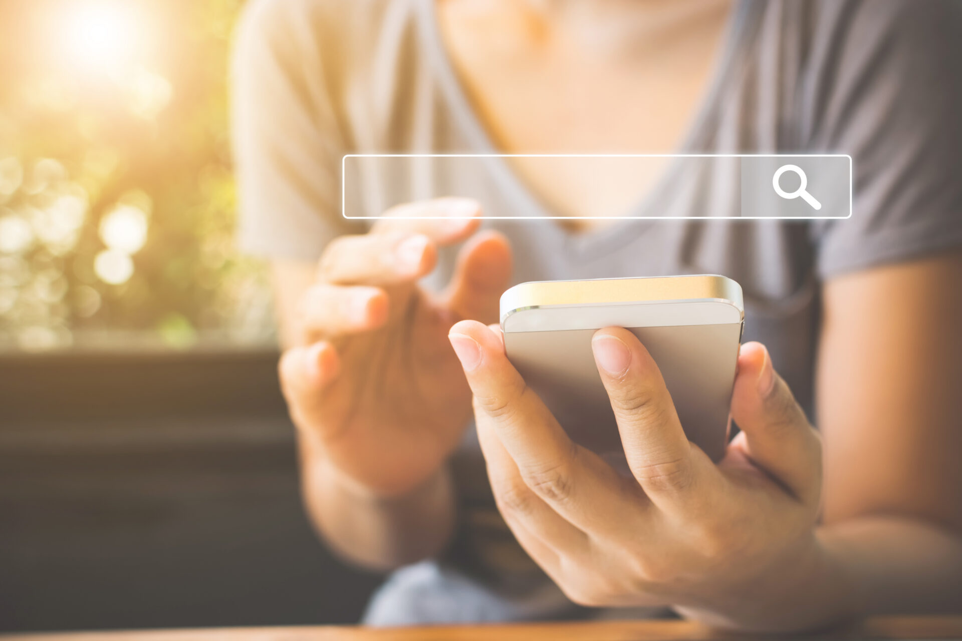 A person uses a smartphone with one hand, touching the screen with the other. A search bar overlays the image as sunlight filters in.