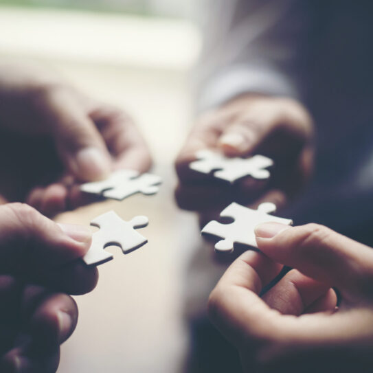 Close-up of four hands holding puzzle pieces, positioned to connect them, representing teamwork, collaboration, and the 4 pillars of a marketing partnership.