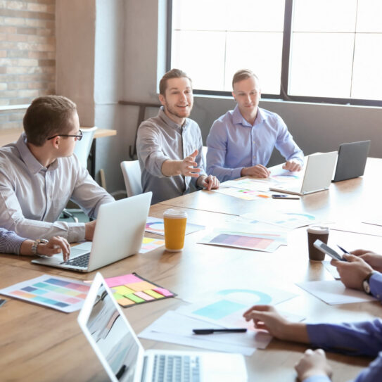 Six people sit at a conference table with laptops and papers. One man gestures as he speaks; others listen in a bright, modern office.