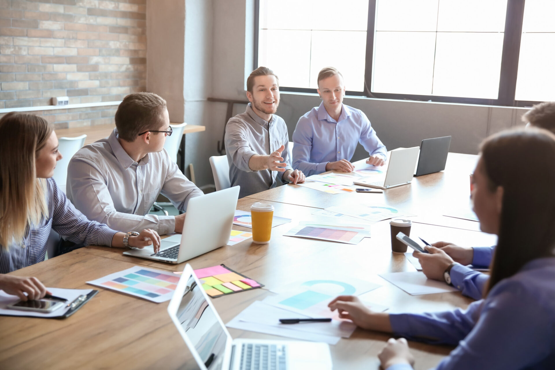 Six people sit at a conference table with laptops and papers. One man gestures as he speaks; others listen in a bright, modern office.