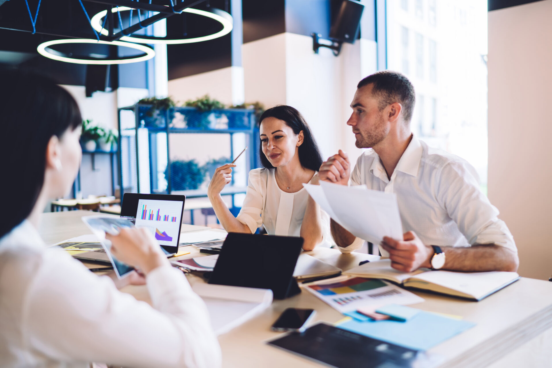 Three people at a modern office table discuss documents and review charts, with papers and devices on the table, plants in the background.