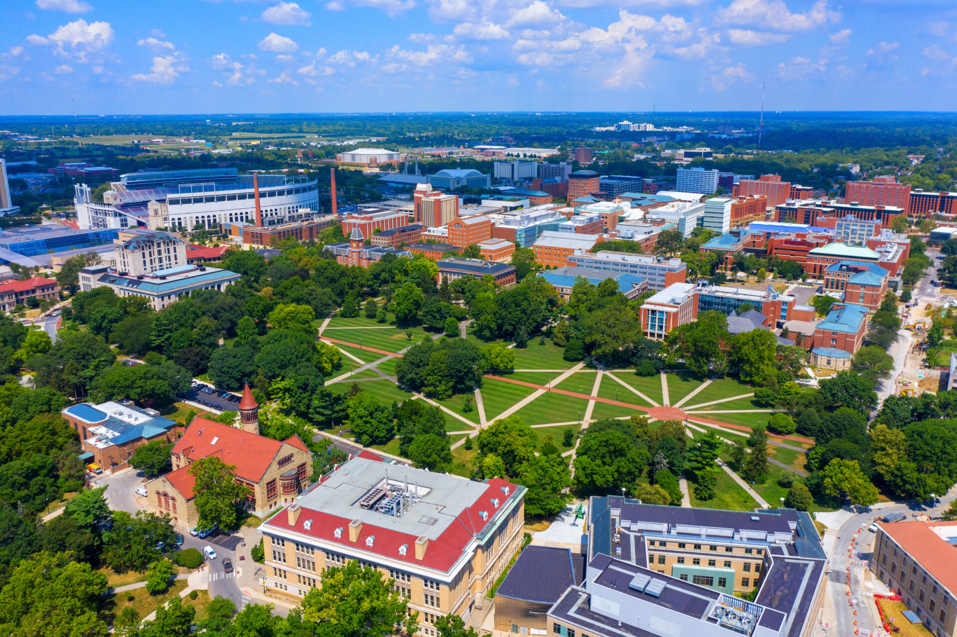 Aerial view of a university campus with red-roofed buildings, green lawns, geometric tree-lined paths, and a stadium under clouds.
