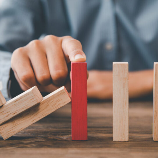 A hand blocks falling wooden dominoes using a single red block, representing intervention or stopping a chain reaction.
