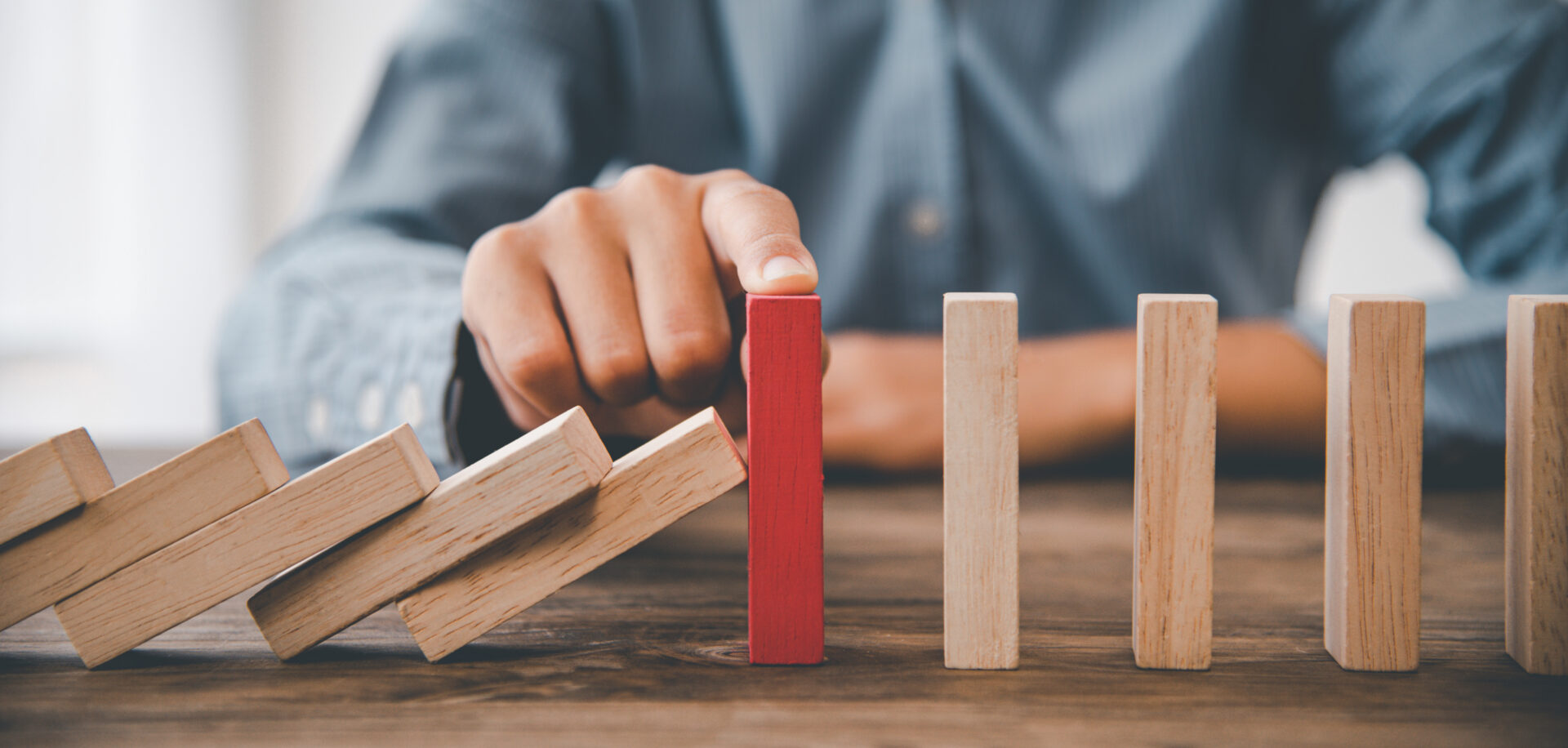 A hand blocks falling wooden dominoes using a single red block, representing intervention or stopping a chain reaction.