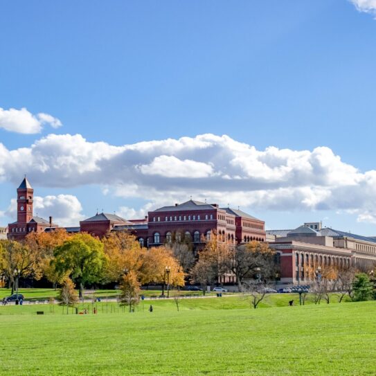 A wide grassy lawn with autumn-colored trees and several large historic red-brick buildings under a blue sky with scattered clouds.