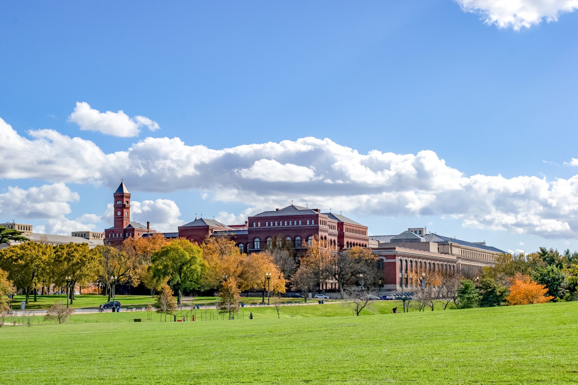 A wide grassy lawn with autumn-colored trees and several large historic red-brick buildings under a blue sky with scattered clouds.