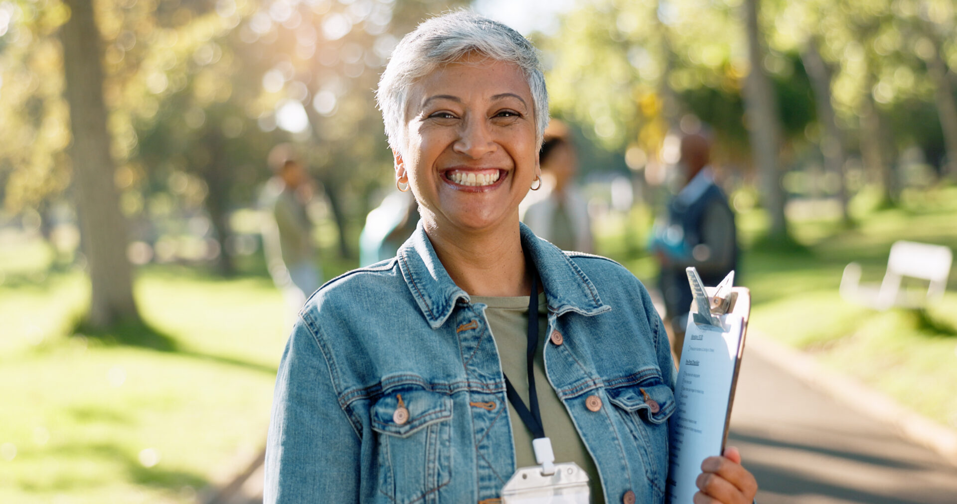 A smiling woman with short gray hair, wearing a denim jacket and holding a clipboard, stands outdoors in a sunlit park.