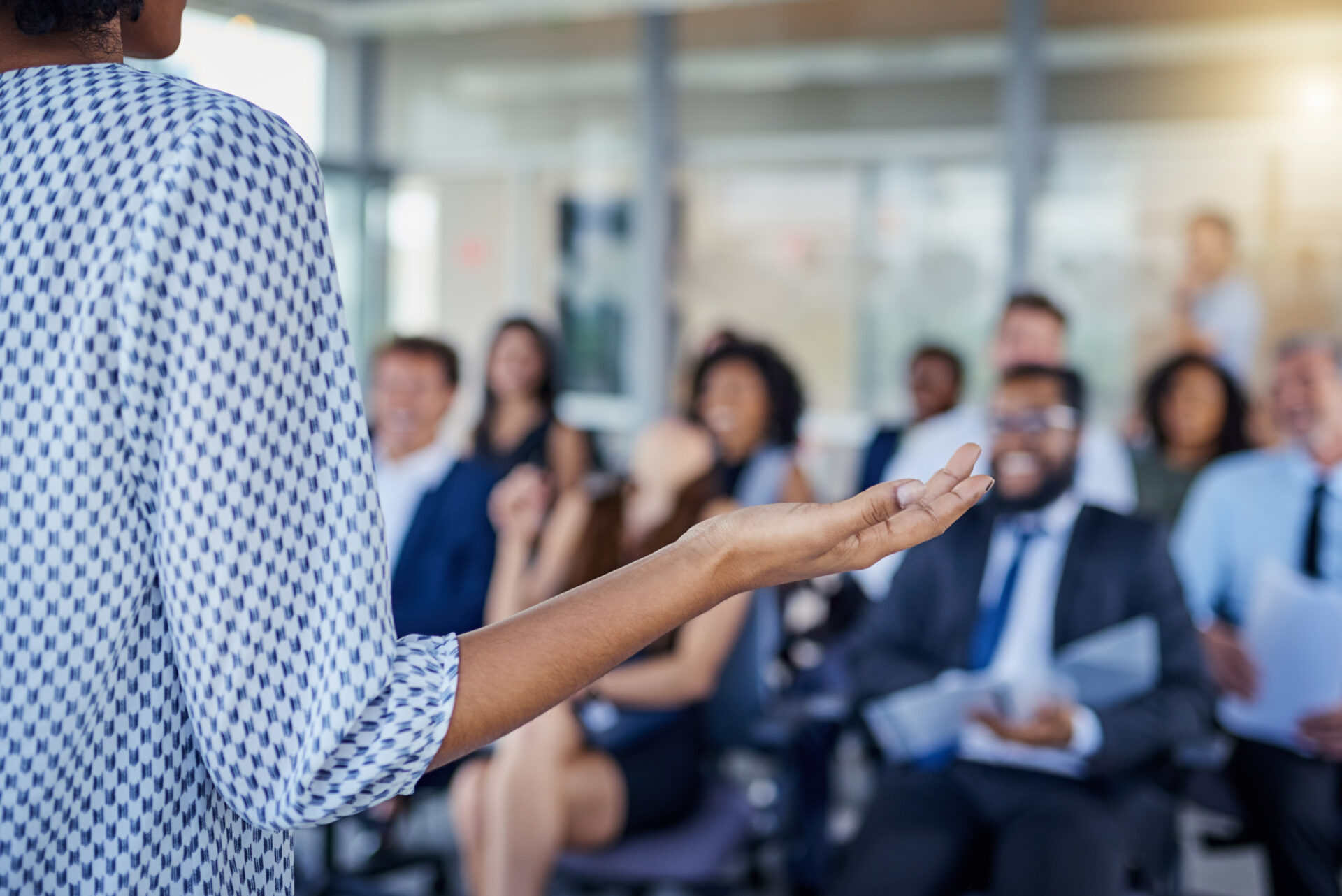 A person gestures with their hand while speaking to a smiling, seated group in a modern office, suggesting an engaging presentation.