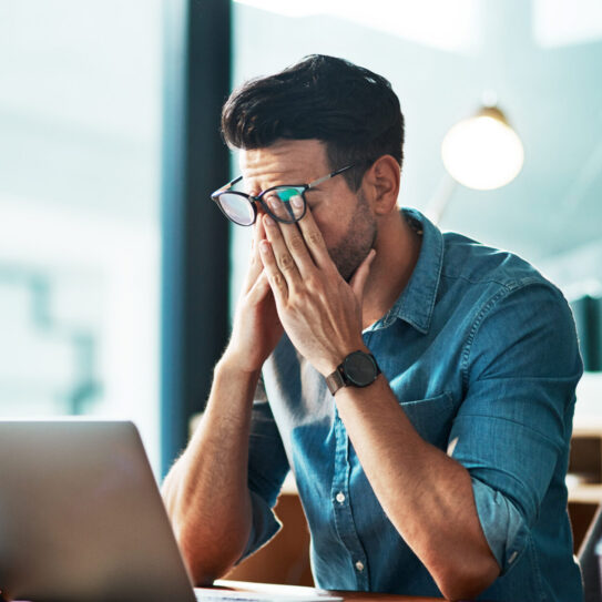 A man in glasses and a denim shirt sits at a desk, rubbing his eyes in stress before an open laptop, with blurred office lights behind.